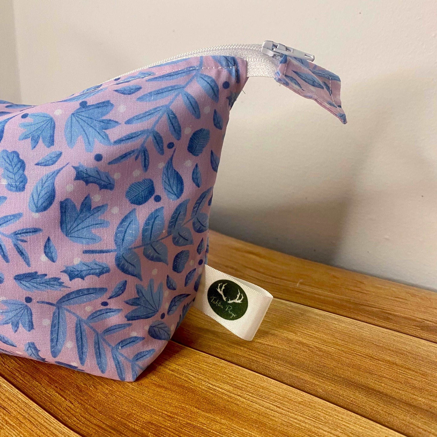Closeup detail of the makeup bag, showing the white zip on the top and the Tahlia Paige label on the side. It is blue leaves on a pink background for the pattern, and it sits on a wooden surface