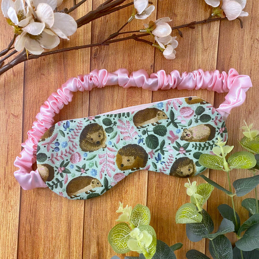 hedgehog patterned sleepmask on a wooden surface with foliage and flowers around it. The pattern is mostly green with the brown hedgehogs and plenty of little pink leaves and flowers.