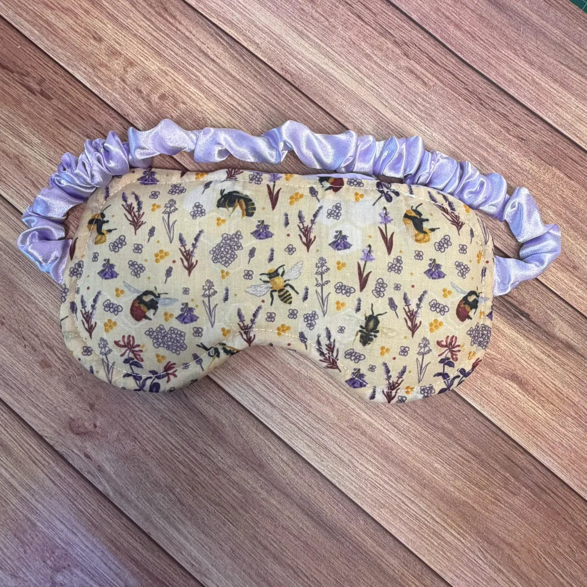 A sleep mask with a light purple satin ruffled strap, featuring a cream-colored printed fabric with a floral and bee pattern, laid flat on a wooden surface.