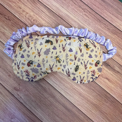 A sleep mask with a light purple satin ruffled strap, featuring a cream-colored printed fabric with a floral and bee pattern, laid flat on a wooden surface.