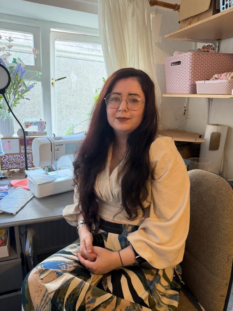 Tahlia Paige, welsh artist, sits in a sewing room, wearing a cream blouse and a patterned skirt. A sewing machine is on the desk in front of her, with shelves and storage boxes in the background.