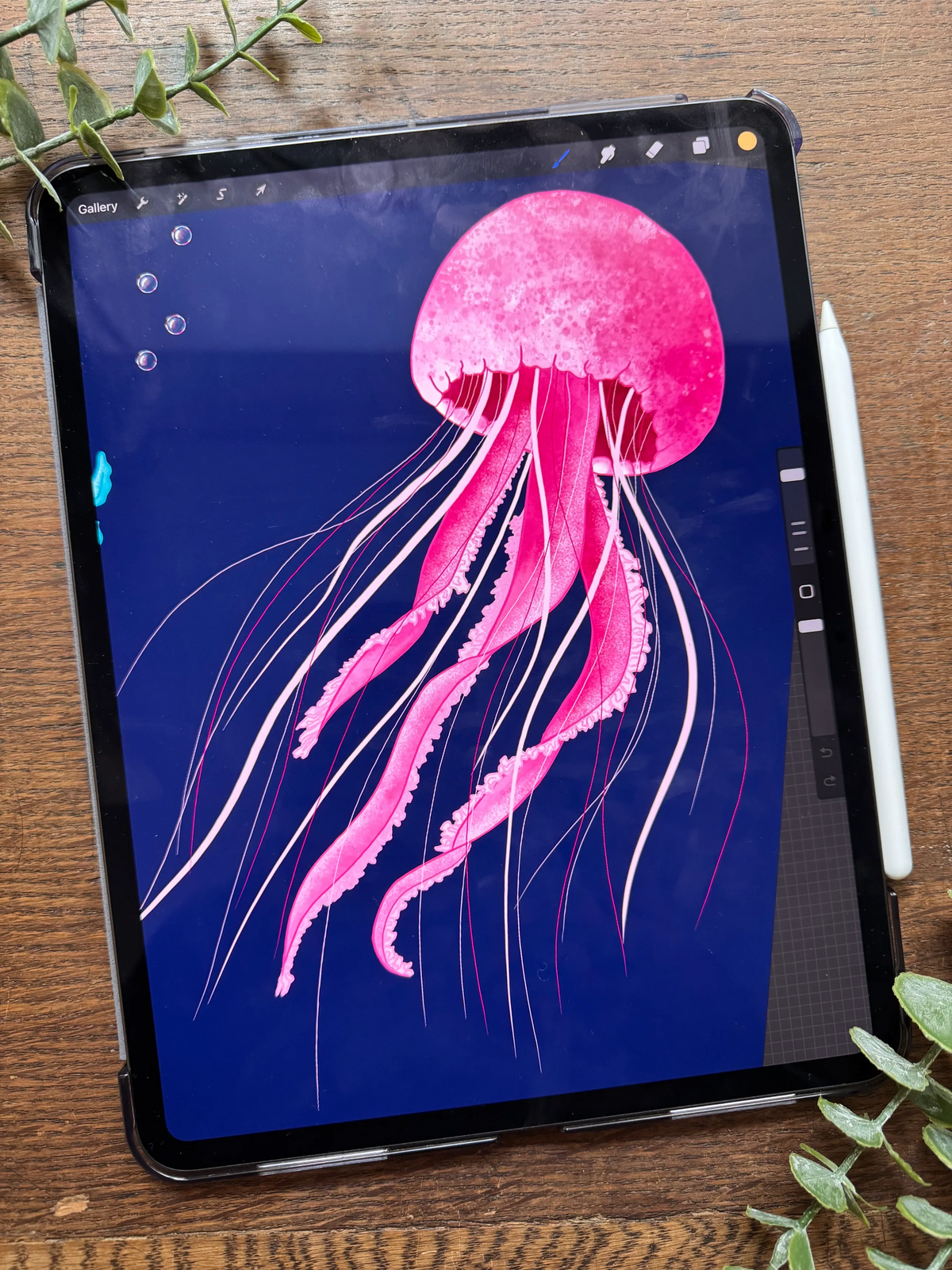 A black tablet on a wooden surface displaying a pink jellyfish illustration on its screen by welsh artist Tahlia Paige, with a white stylus resting beside it and small green leaves around the edges.