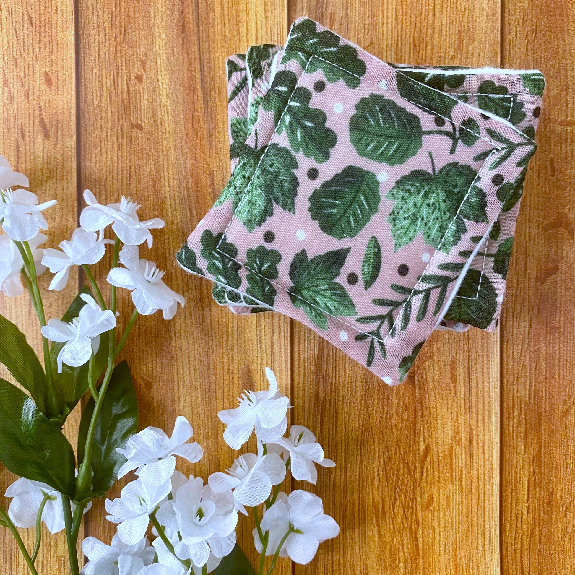 A stack of green foliage patterned skincare pads sat on a wooden background with some flowers around it
