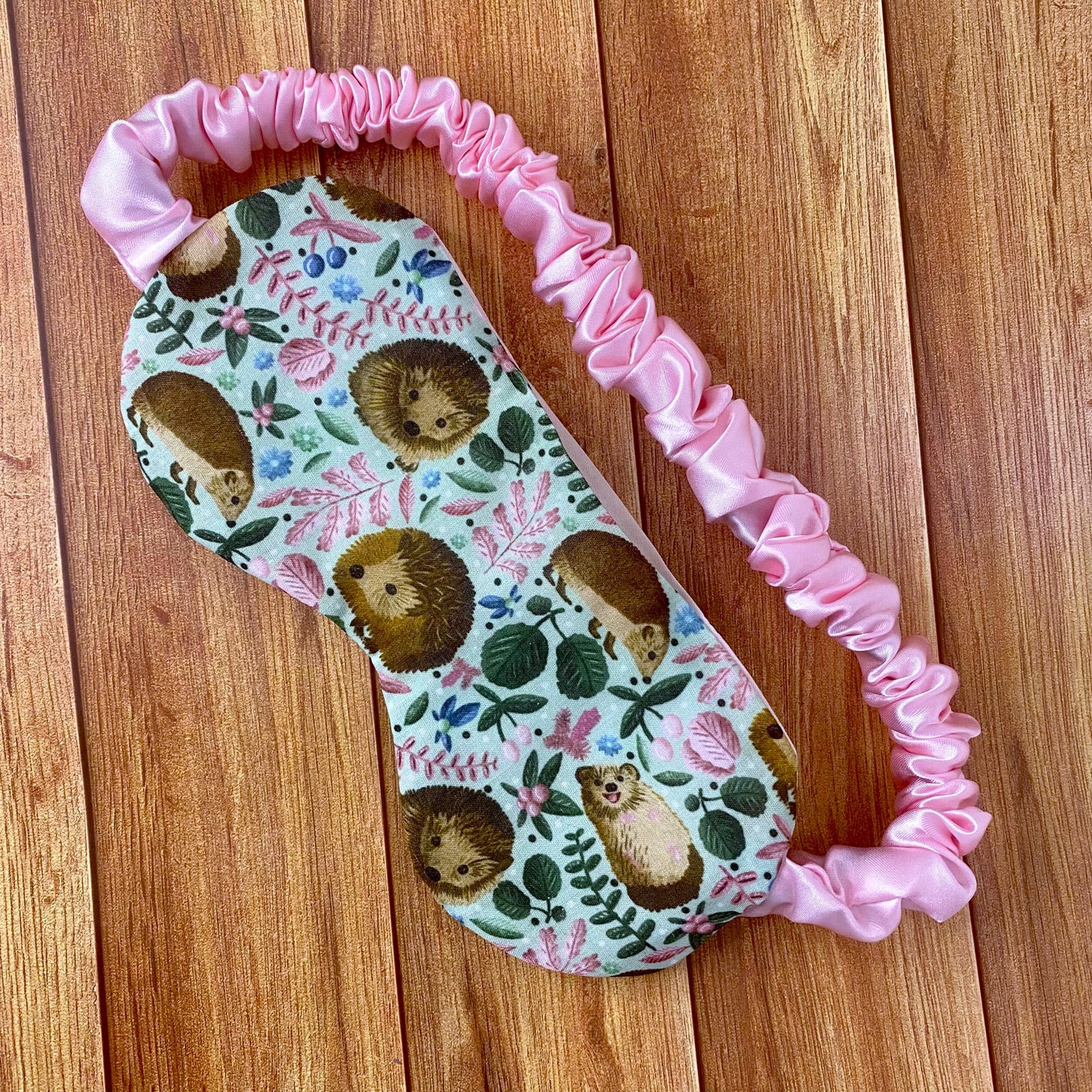 hedgehog patterned sleepmask with a pink scrunched satin strap on a wooden backdrop
