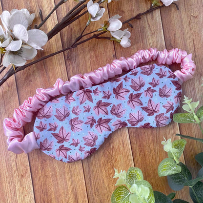pink leafy patterned sleepmask on wooden backdrop with foliage around it. The pattern has illustrated pink leaves on a blue background