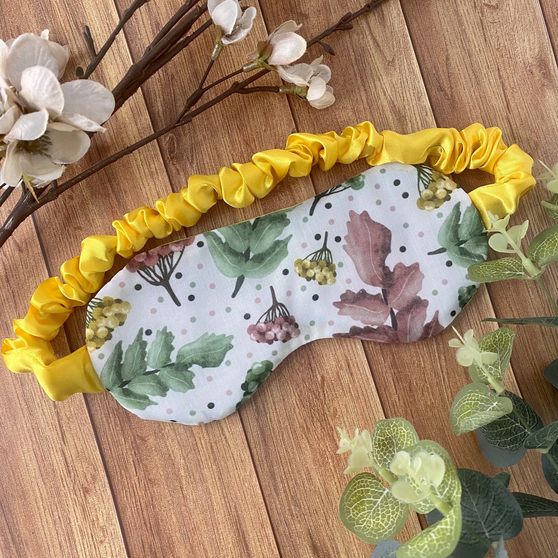 pretty foliage patterned sleepmask with scrunched satin strap on wooden background with flowers and foliage around it
