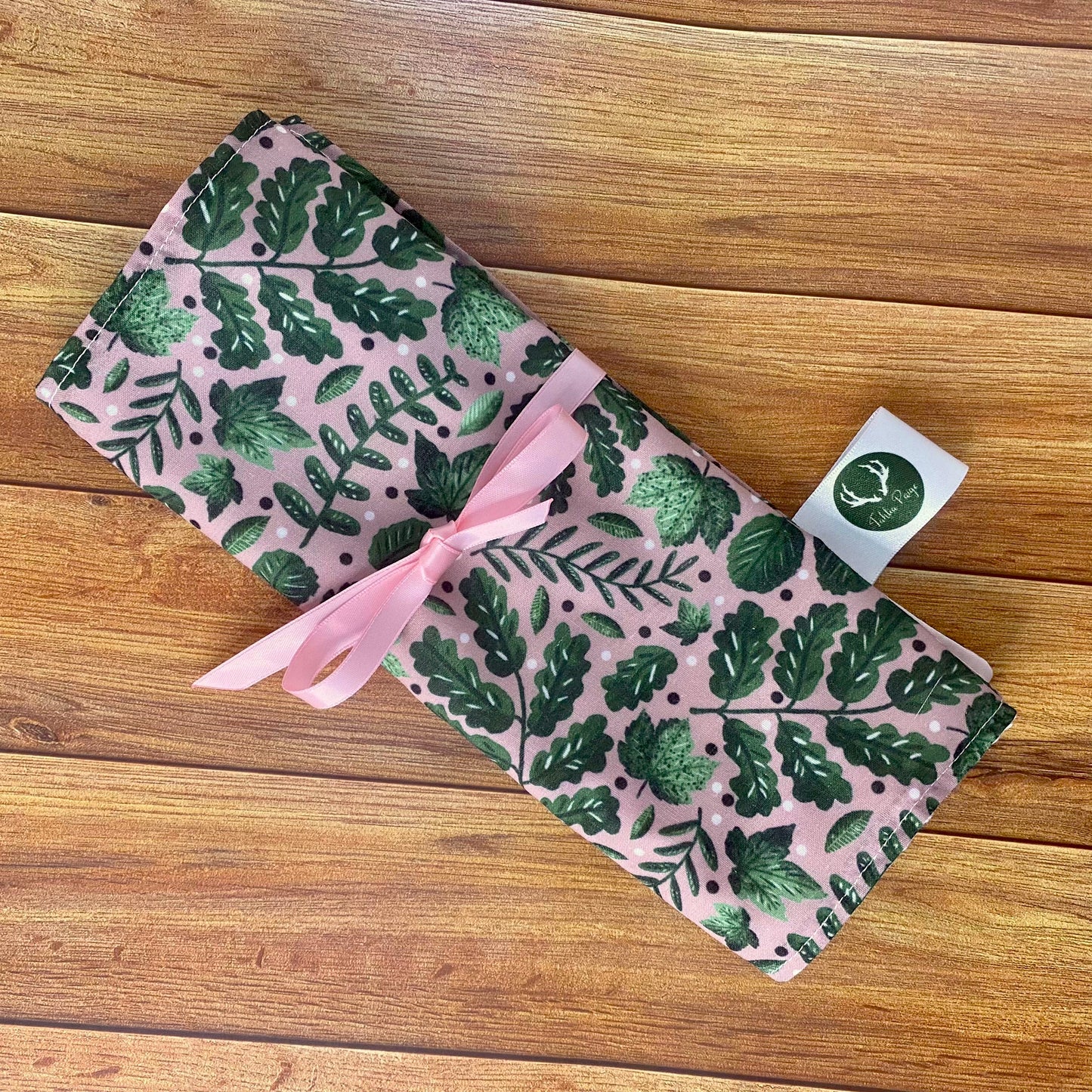 Green foliage brushwrap sat on a wooden background all folded up and sealed shut with a pink ribbon. The surface pattern design on the front is a green collection of leaves on a pink background.