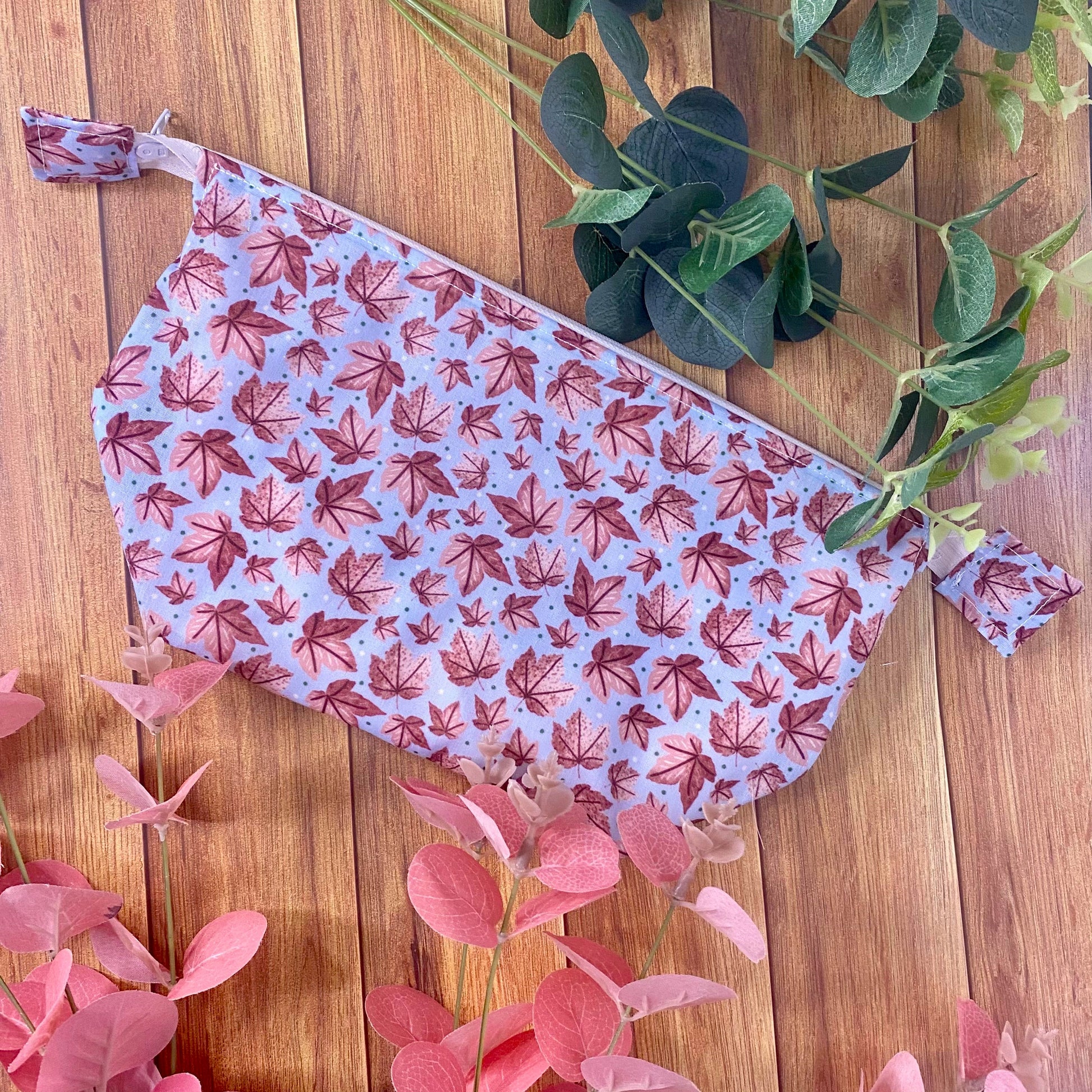 photograph of a brightly patterned makeup bag on a wooden background, with green and pink foliage around it. The surface pattern design includes pink sycamore leaves on a blue background with some dots.