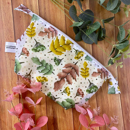 a photograph of a makeup bag flat on a wooden background with foliage around it. The bag is patterned with a white surface pattern design that includes pink, green and yellow berries and leaves and spots.