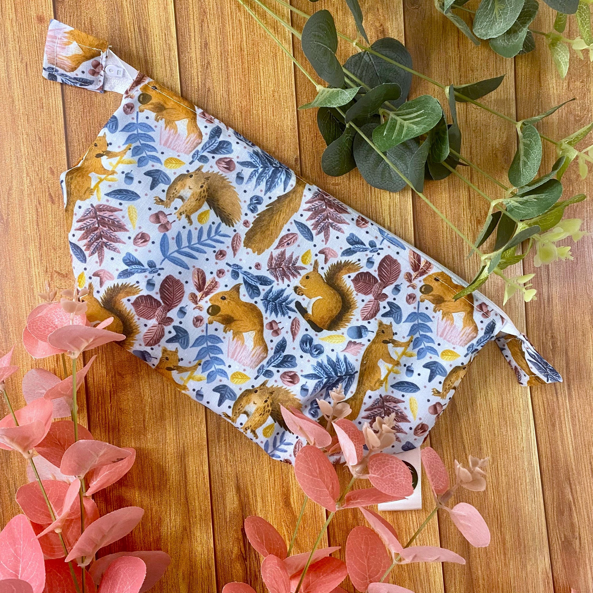 a makeup bag sat on the wooden background with green and pink foliage around it. The makeup bag has a red squirrel pattern on it, mostly blue with hints of pink and yellow throughout on small leaves and things.