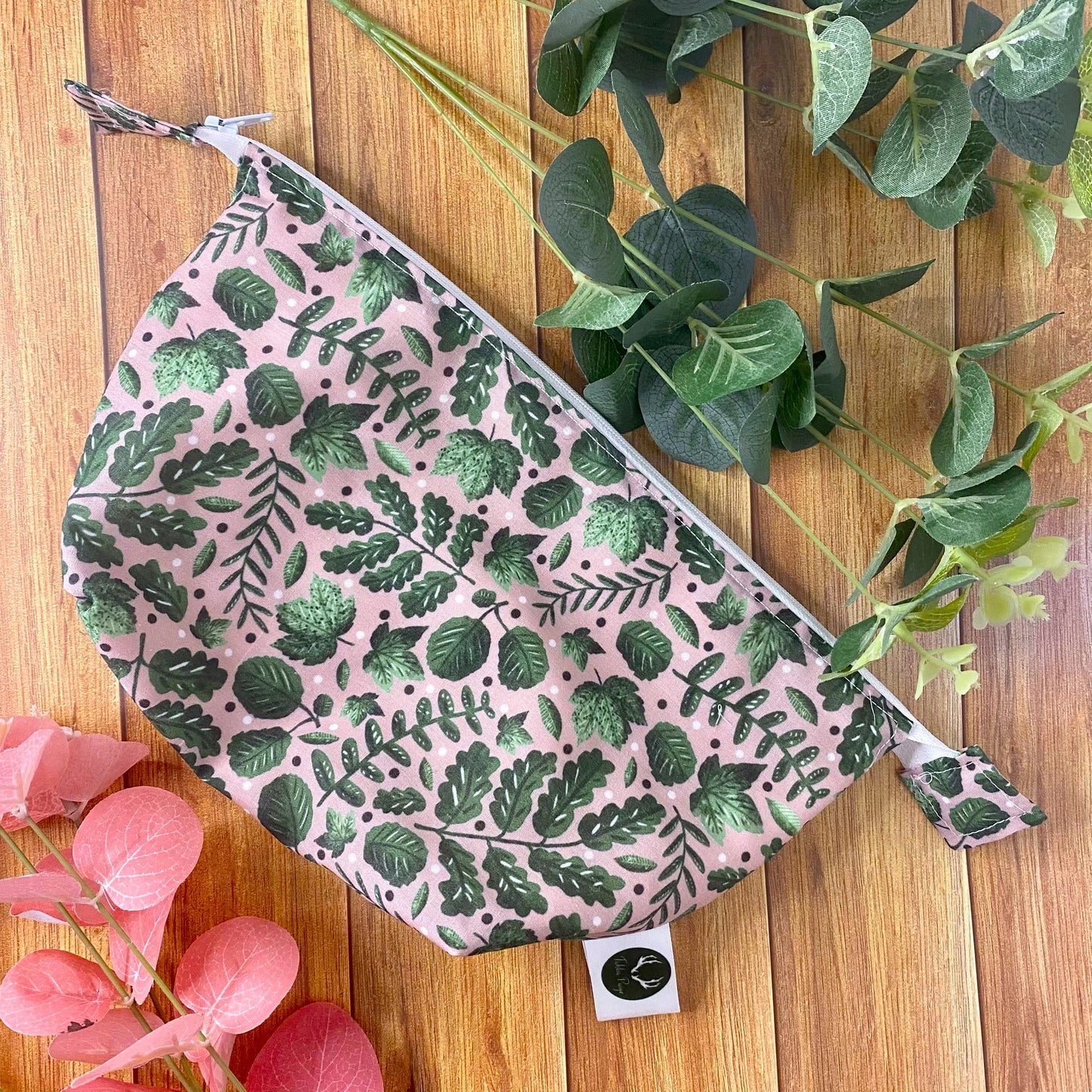 photograph of a green foliage makeup bag on a wooden background with green and pink foliage around it.