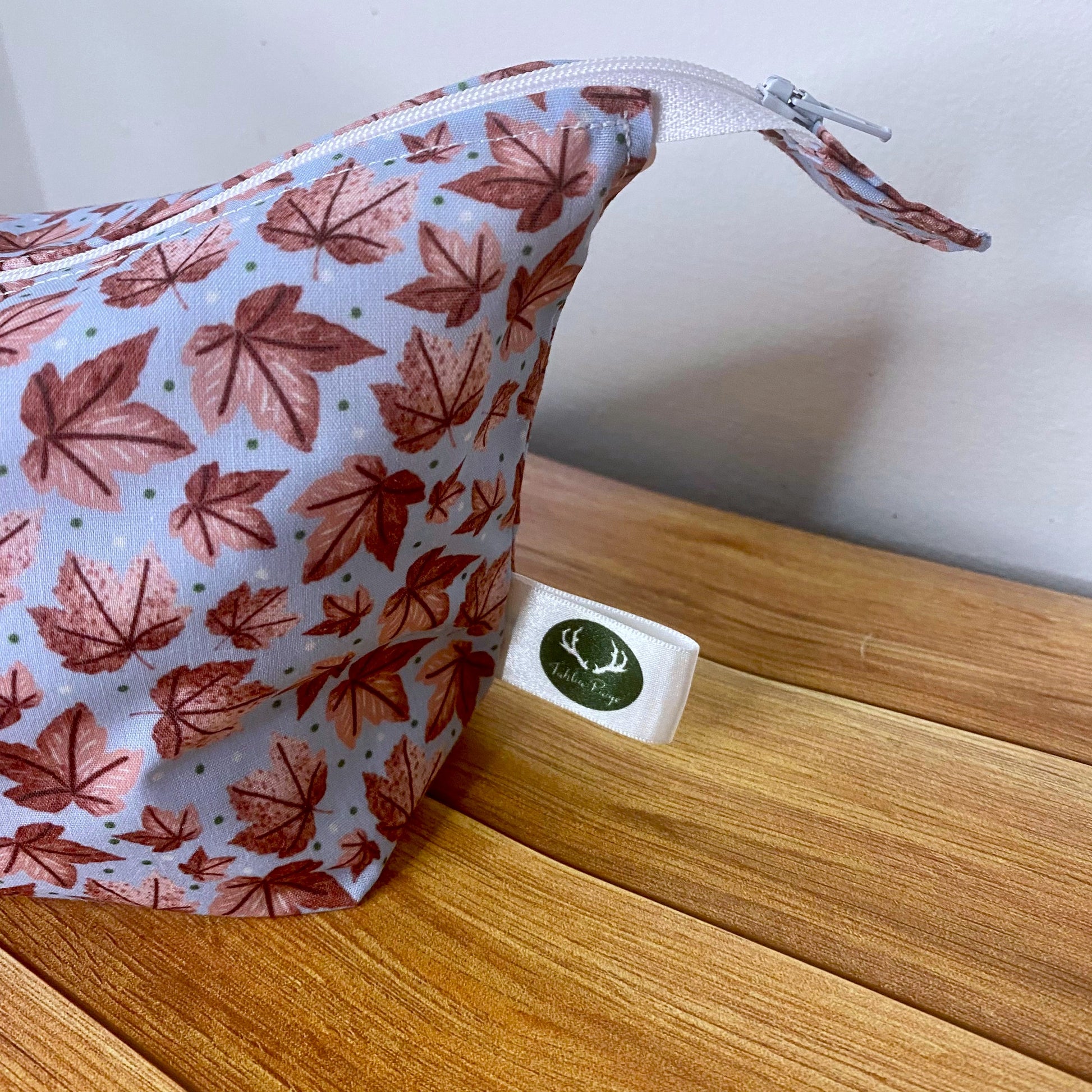 Closeup photograph of the side of the pink leafy patterned makeup bag, the bag is stuffed to show the shape of the side of the bag. The bag has the Tahlia Paige label sticking out the side seam, and it is sat on a wooden background.