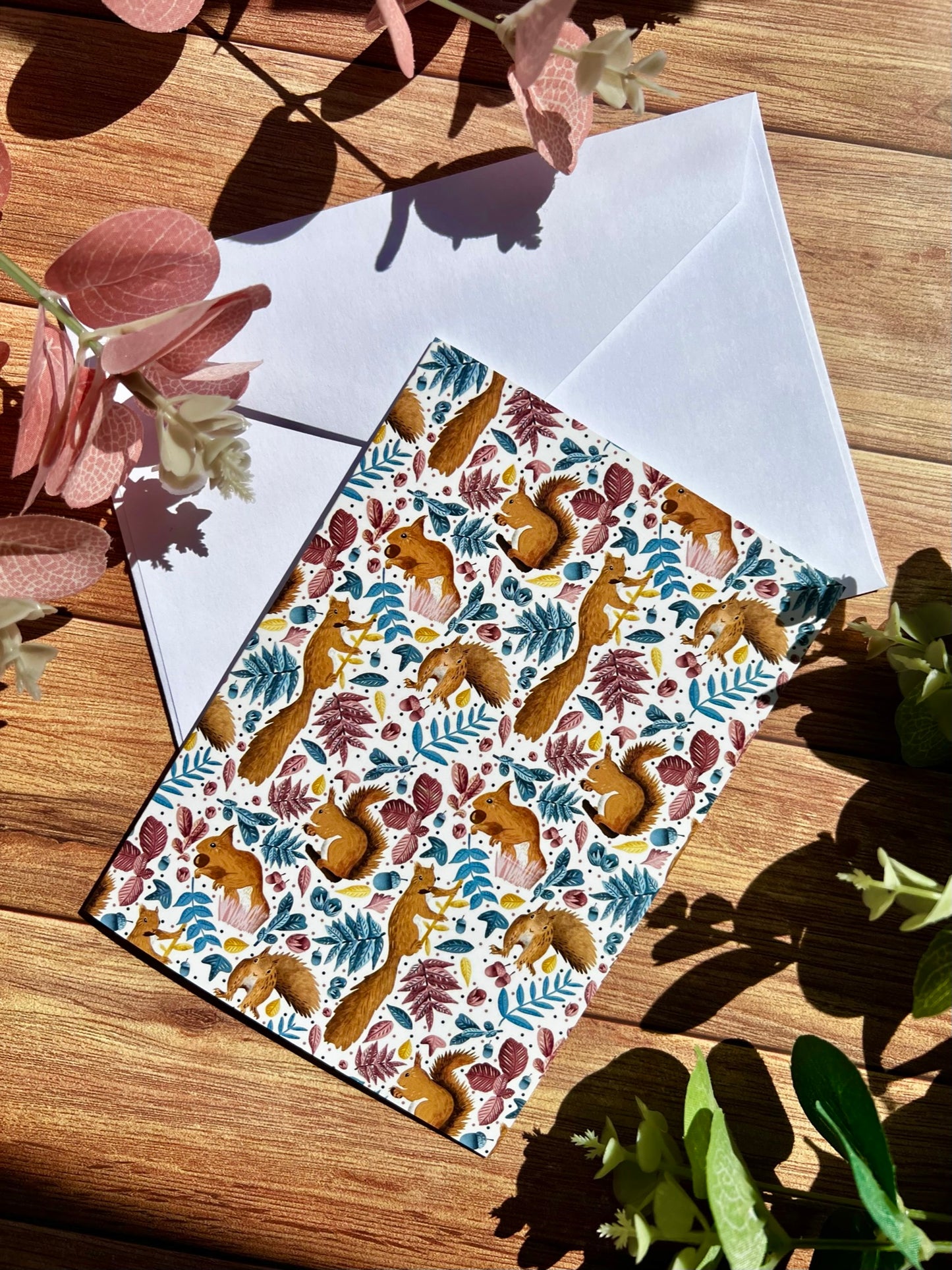 A small greetings card featuring a red squirrel surface pattern design lying on top of an envelope. Both are lying on a wooden surface with pink and green foliage around the card.