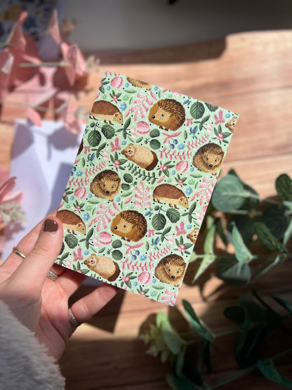 Small greetings card with a green hedgehog surface pattern design on it being held in the sunlight with various green and pink foliage blurred behind it