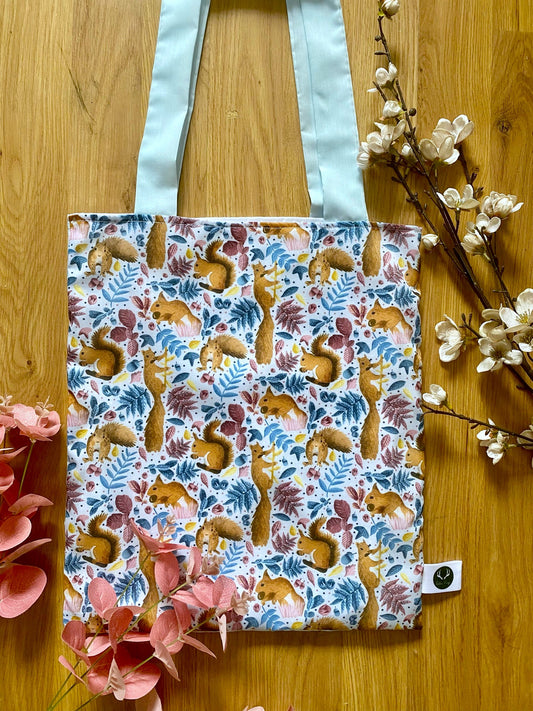 red squirrel tote bag on a wooden backdrop with foliage around it. The bag has blue handles to compliment the pattern, which has blue and pink leaves and things around the little squirrels in the design.