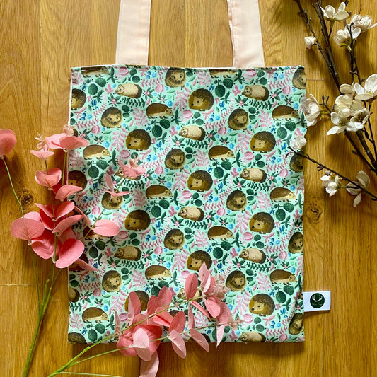 Closeup of the hedgehog pattern on the reusable shopping tote,. It is sat on a wooden background with pink foliage around it.