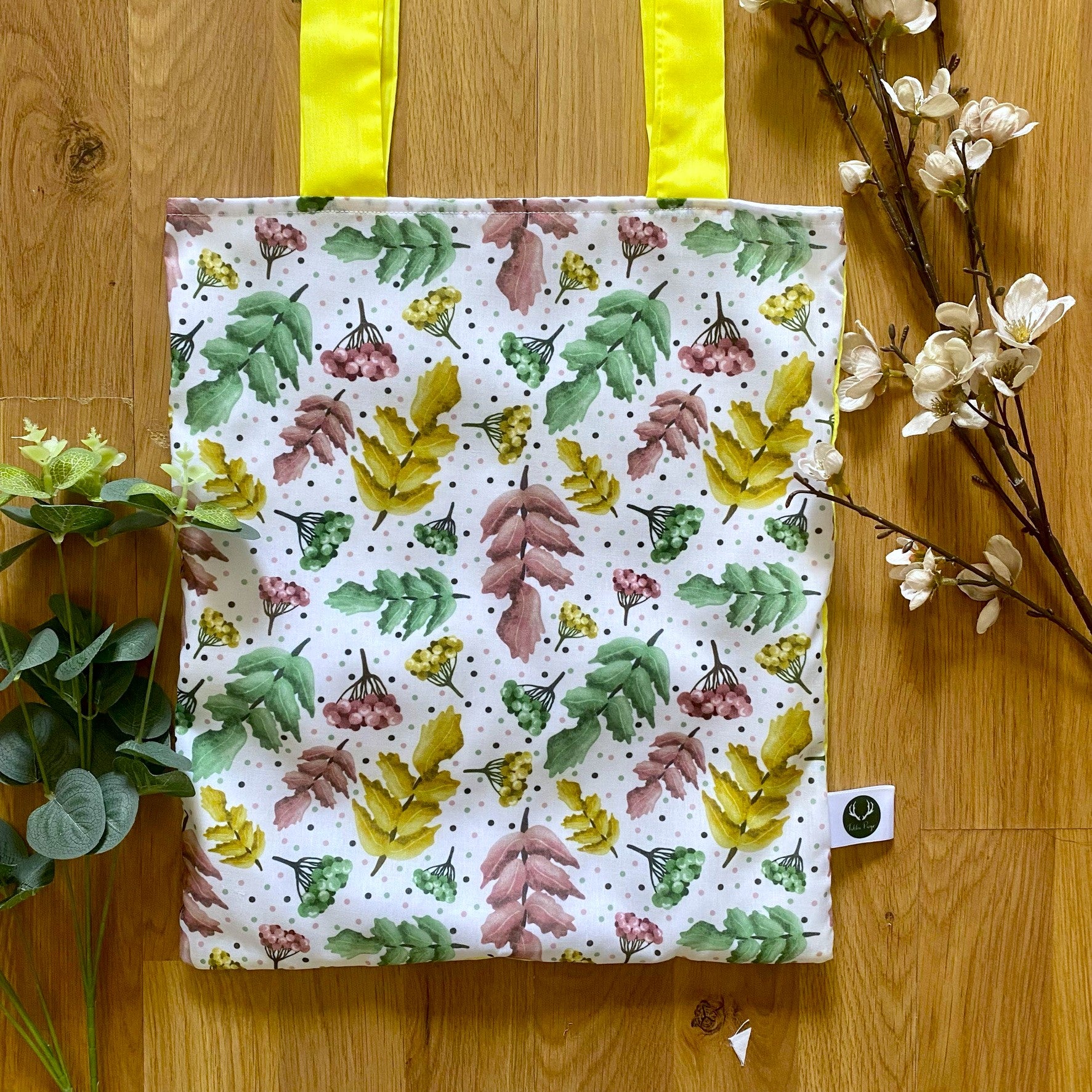 Closeup of surface pattern design on the front of the pretty foliage tote bag, with yellow handles, and sat on a wooden backdrop with foliage around it