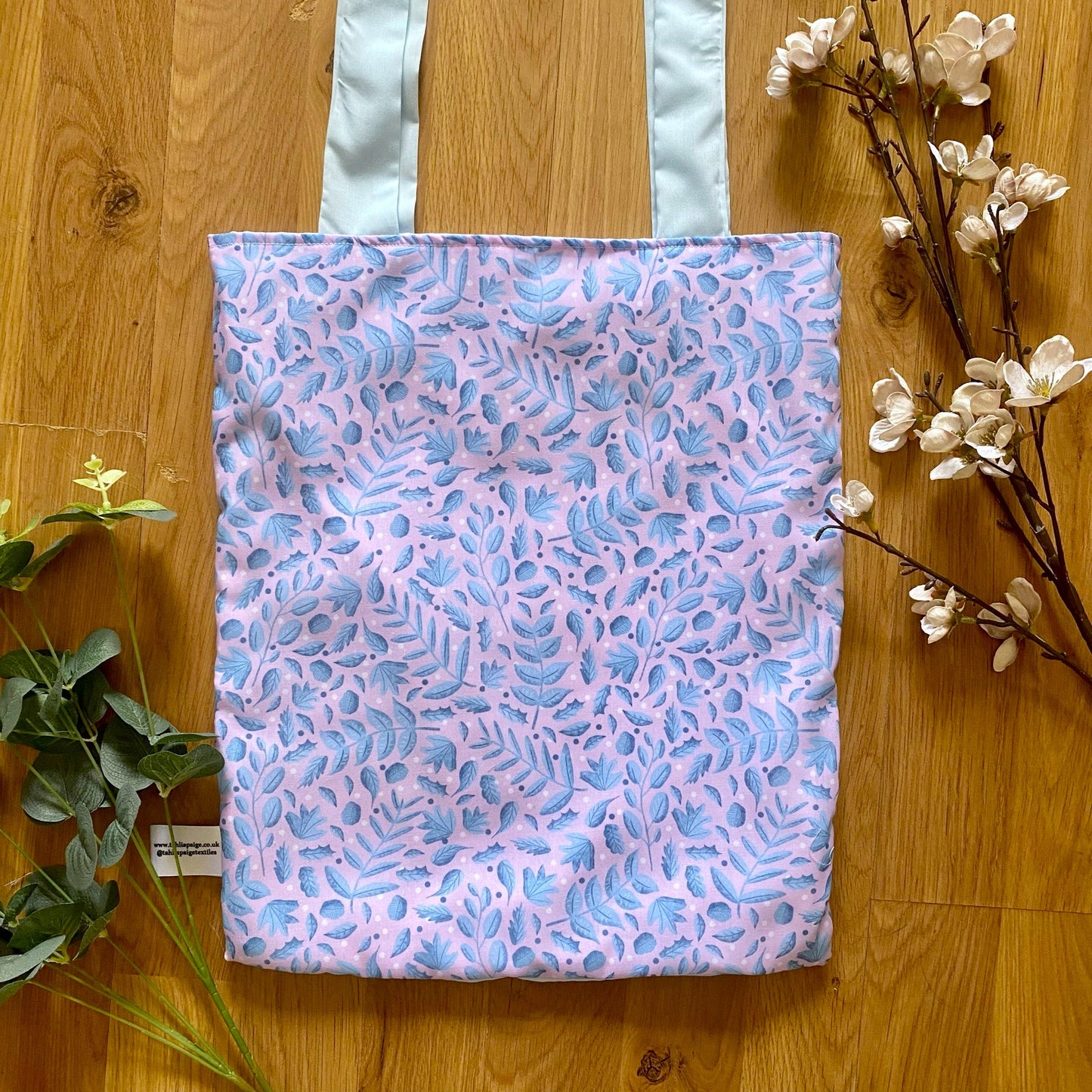 blue and pink foliage shopping bag lying flat on the wooden background with green and pink foliage around it