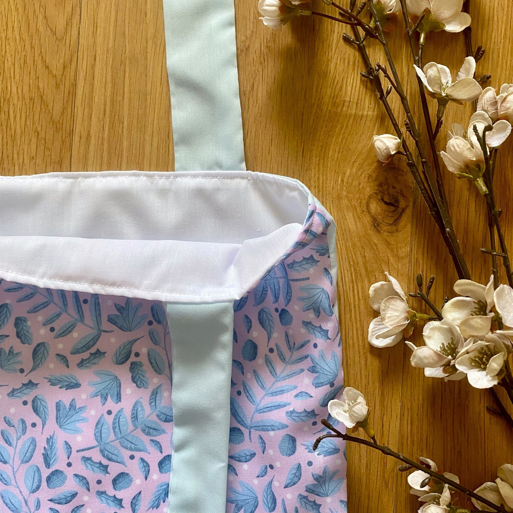 closeup of the blue and pink foliage tote bag with the blue handles and white lining showing, on a wooden background with blossom next to it