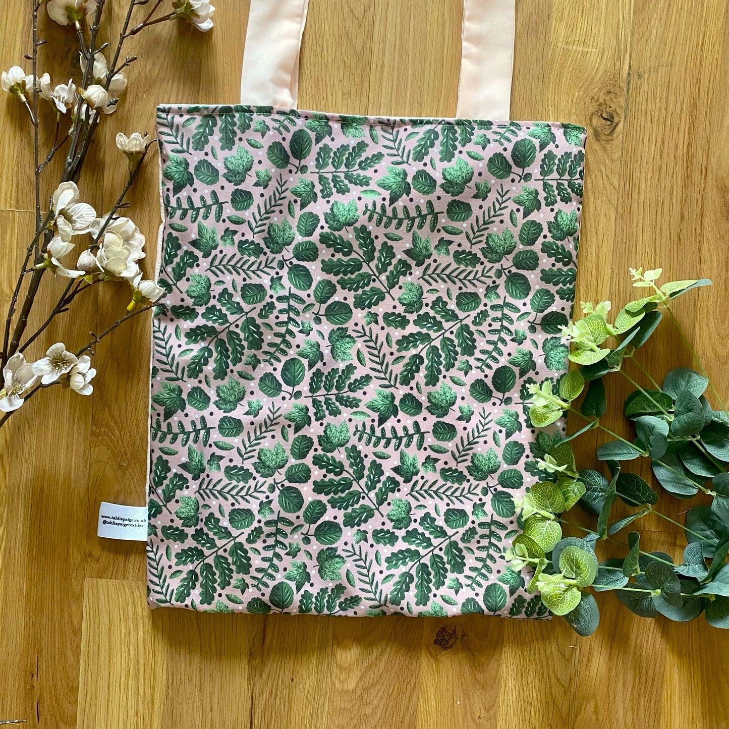 Closeup of the green foliage tote bag on a wooden background with green and pink foliage around it. The green foliage pattern on the bag is green leaves on a pink background.