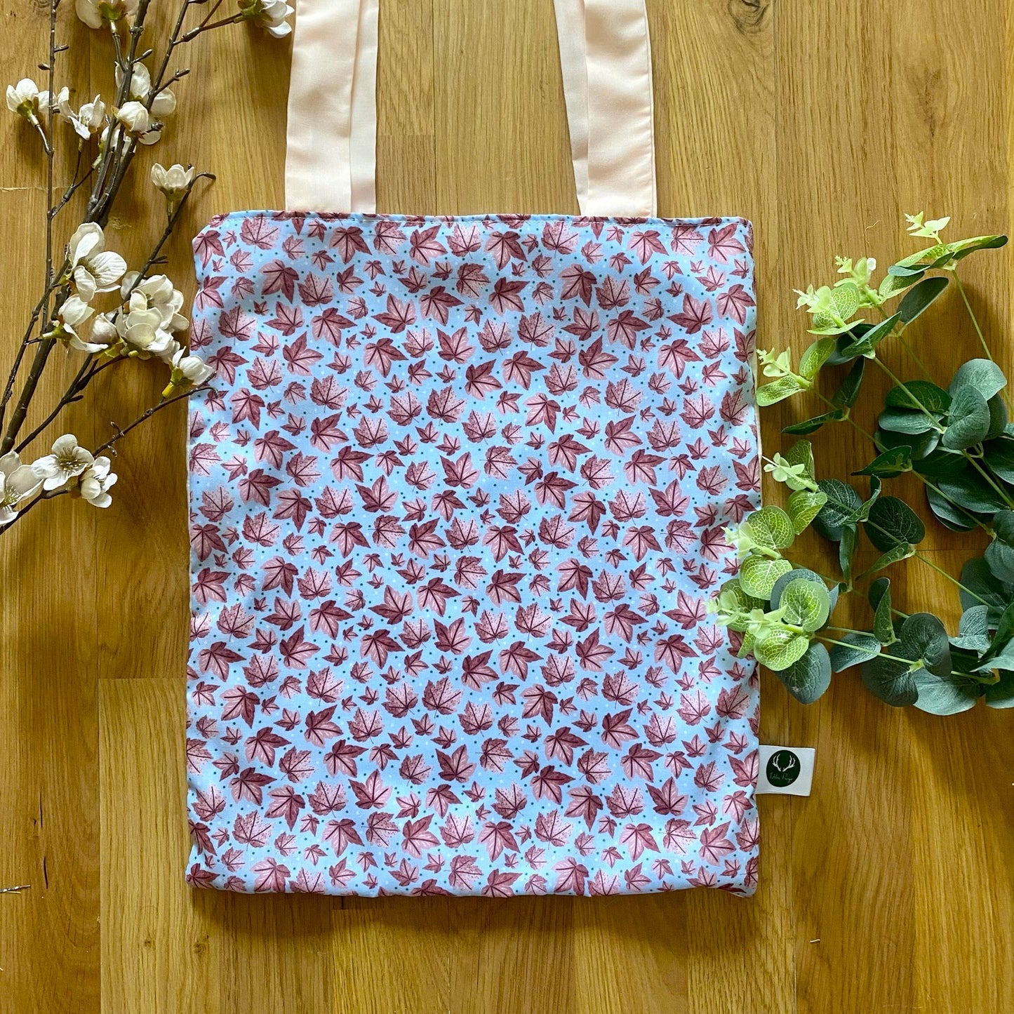 Closeup of surface pattern design on the front of the tote bag, showing a pink leaf pattern with a blue background. The tote bag is shown on a wooden background with green foliage around it.