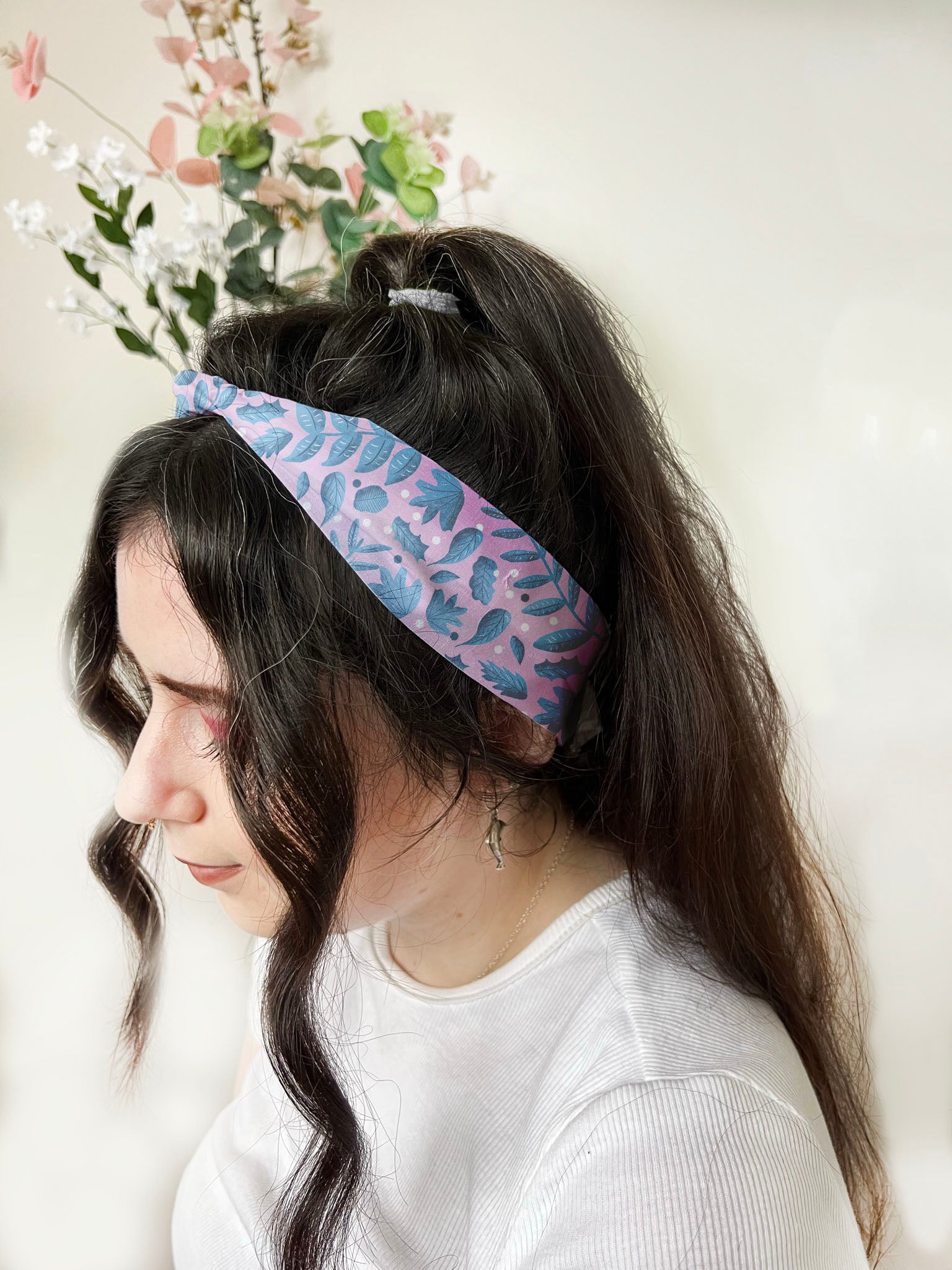 Dark haired girl wears a headband with a ponytail behind her head. The headband has blue foliage leaves illustrated on it with a pink background on the fabric. The headband has a knot at the front.