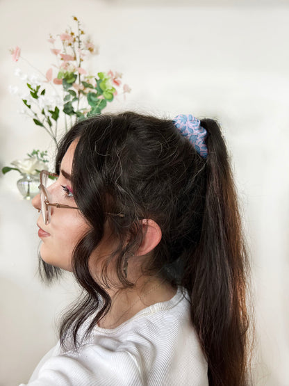 Dark haired girl wearing a scrunchie around a high ponytail, the scrunchie has the blue foliage pattern design on it which is blue illustrated leaves on a pink background