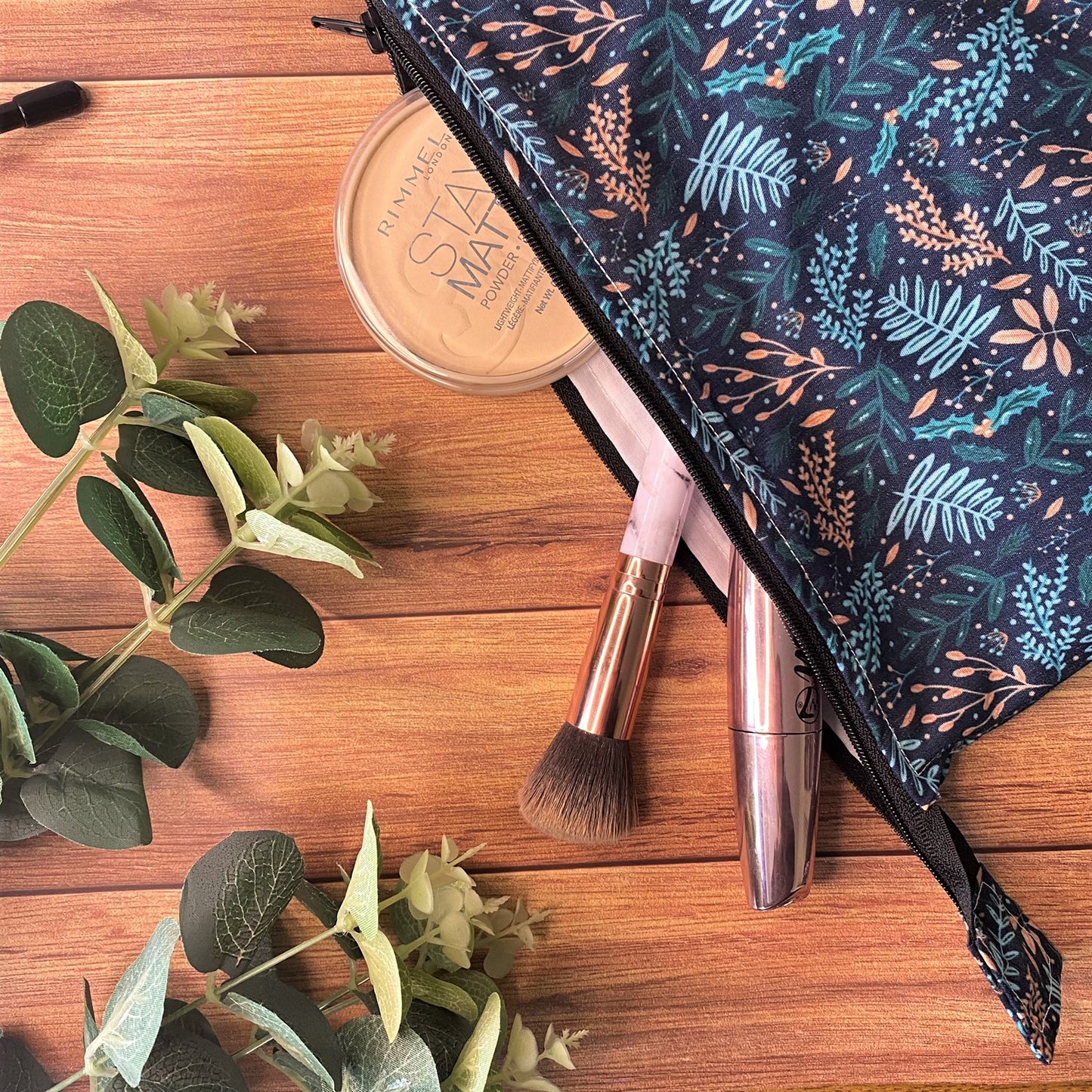 Closeup of the makeup bag with the dark foliage pattern and a makeup brush, mascara and a powder sticking out of the bag to show the size. It is sat on a wooden background near some green foliage
