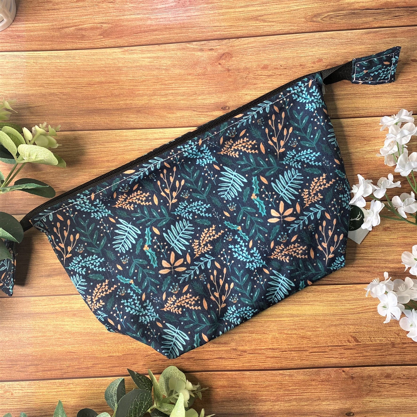dark foliage patterned makeup bag on a wooden background with green and white foliage around it