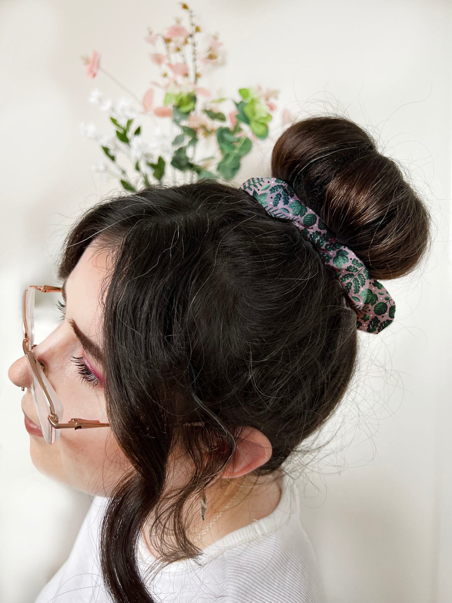Girl with dark hair photographed with up-do bun with a green foliage patterned scrunchie securing it. Plants in background in white room.