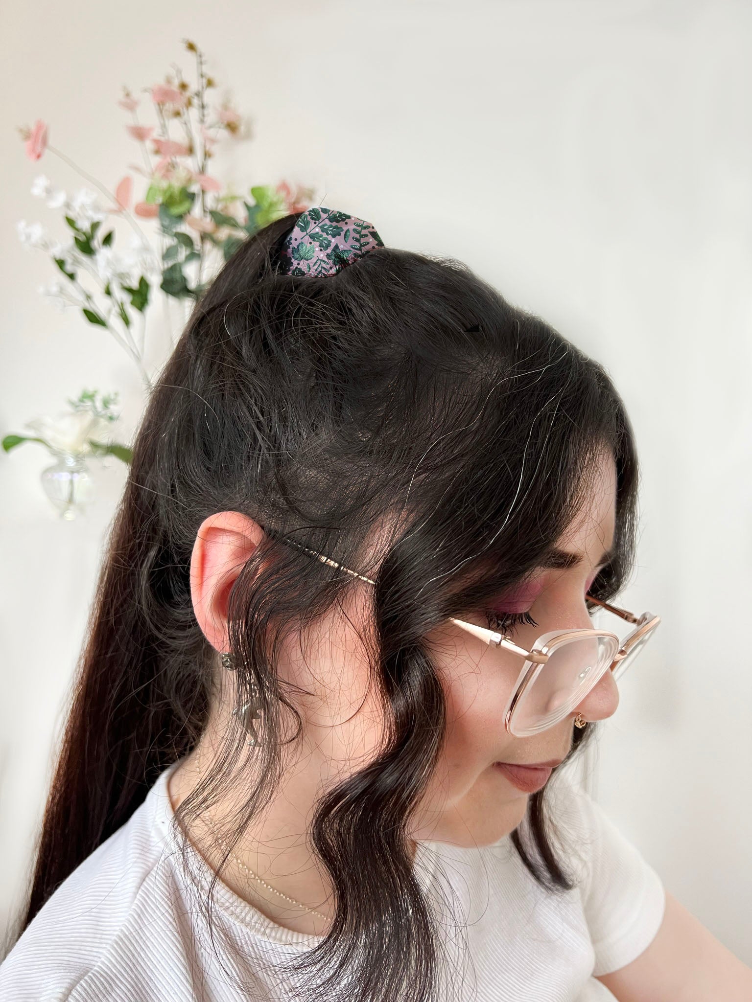 dark haired girl with glasses and ponytail with a scrunchie securing it.  The scrunchie has a green leafy pattern illustrated on it, with a pink background.