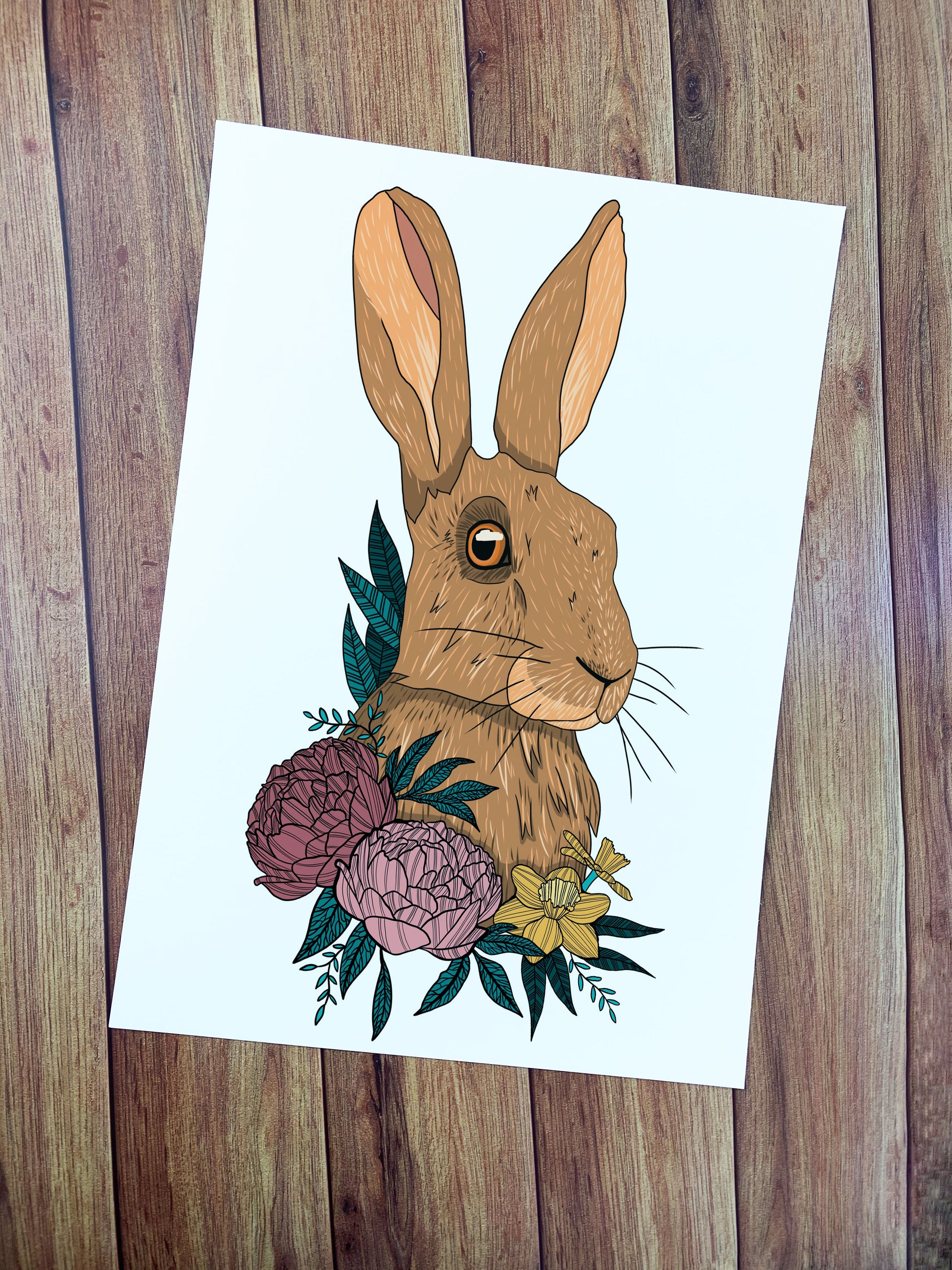 The hare print here shows some yellow daffodils and large pink flowers at the base of a hare's portrait, illustrated in a digital style on a white background. The print then sits on a wooden background.