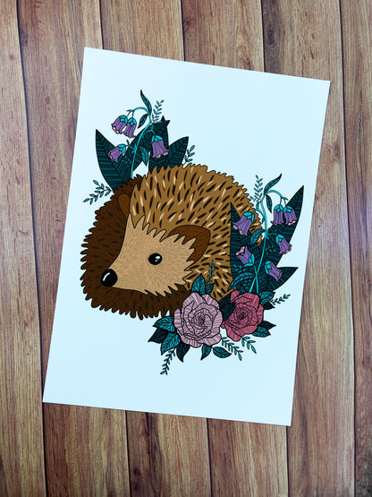 Hedgehog art print shown on a wooden background. The print is an illustration of a happy hedgehog amongst flowers and leaves on a white background.