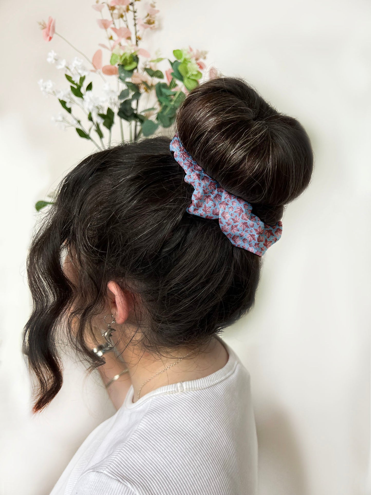dark haired girl wears pink leafy patterned scrunchie in hair around a bun. with plants in the background in a white room