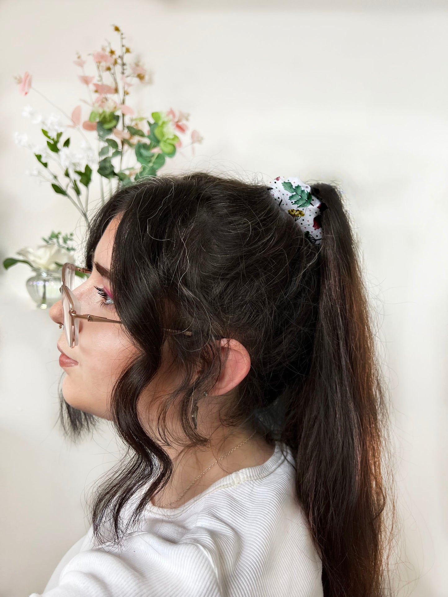 pretty foliage patterned scrunchie in hair of dark haired girl with glasses, facing sideways in white room with plants behind her