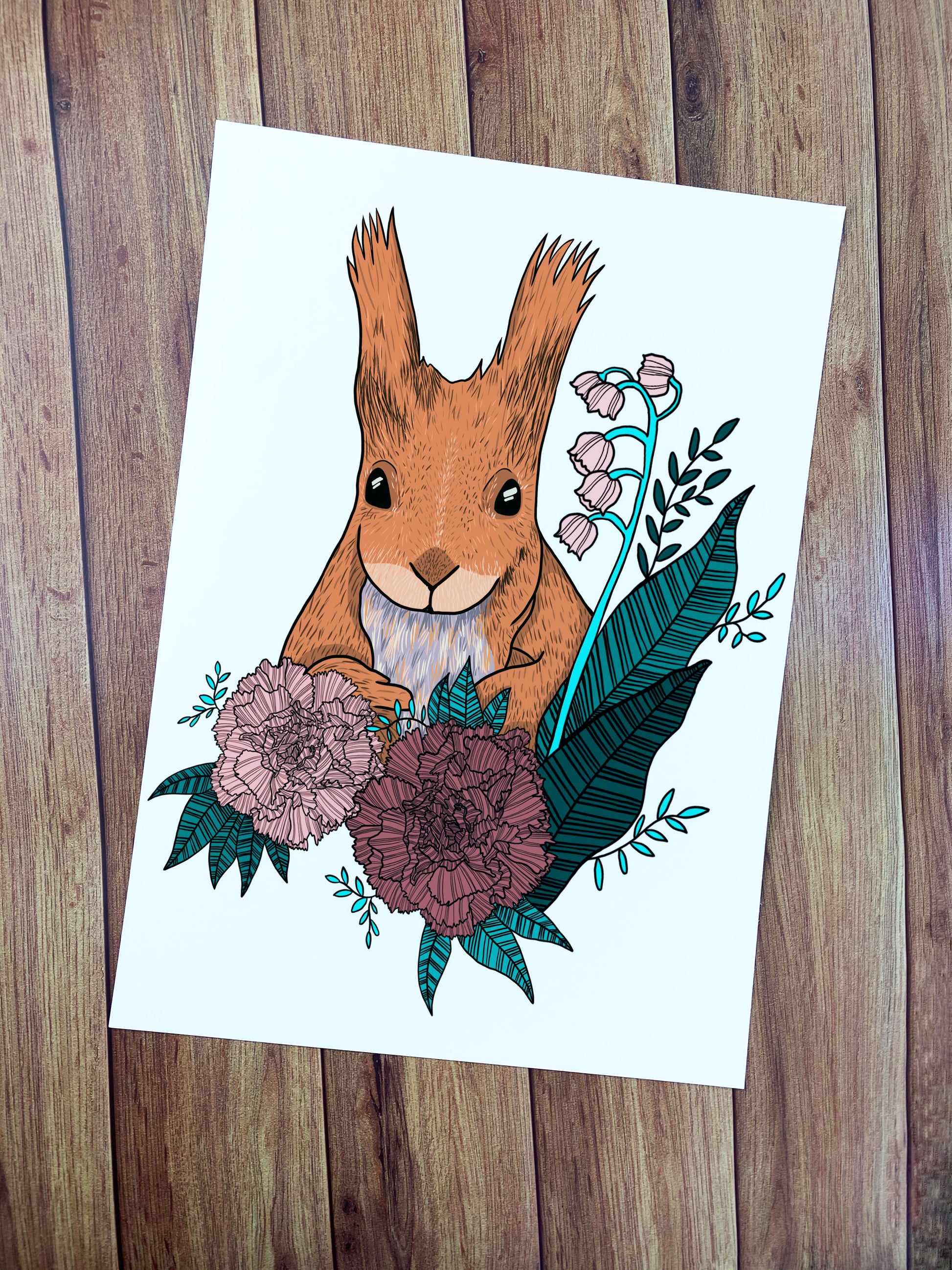 Art print against a wooden background with a red squirrel illustration on it, the squirrel is drawn digitially and sat amongst flowers and leaves. An ideal gift for a red squirrel lover, or someone who cares about endangered species.