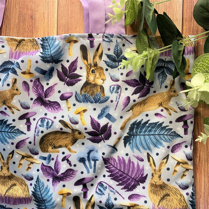Another corner of the hare tote bag with the surface pattern design on show, there is also green foliage over the tote and it is on a wooden backdrop.