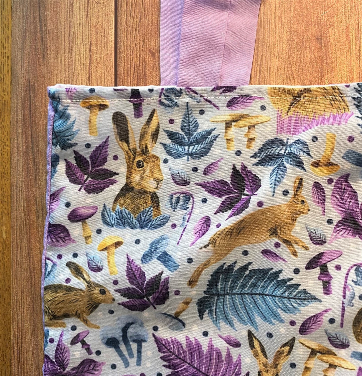 Closeup of the corner of the hare tote showing the surface pattern design with hares and foliage in purple and blue. The purple strap of the tote is also visible and the bag sits on a wooden background.