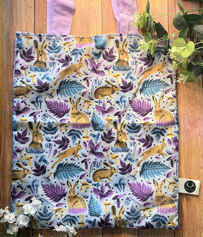 Closeup of pattern on the front of the tote bag which shows hares in a purple and blue surface pattern design. The design features a range of blue and purple leaves, flowers and mushrooms, and covers the whole of the front of the tote bag.
