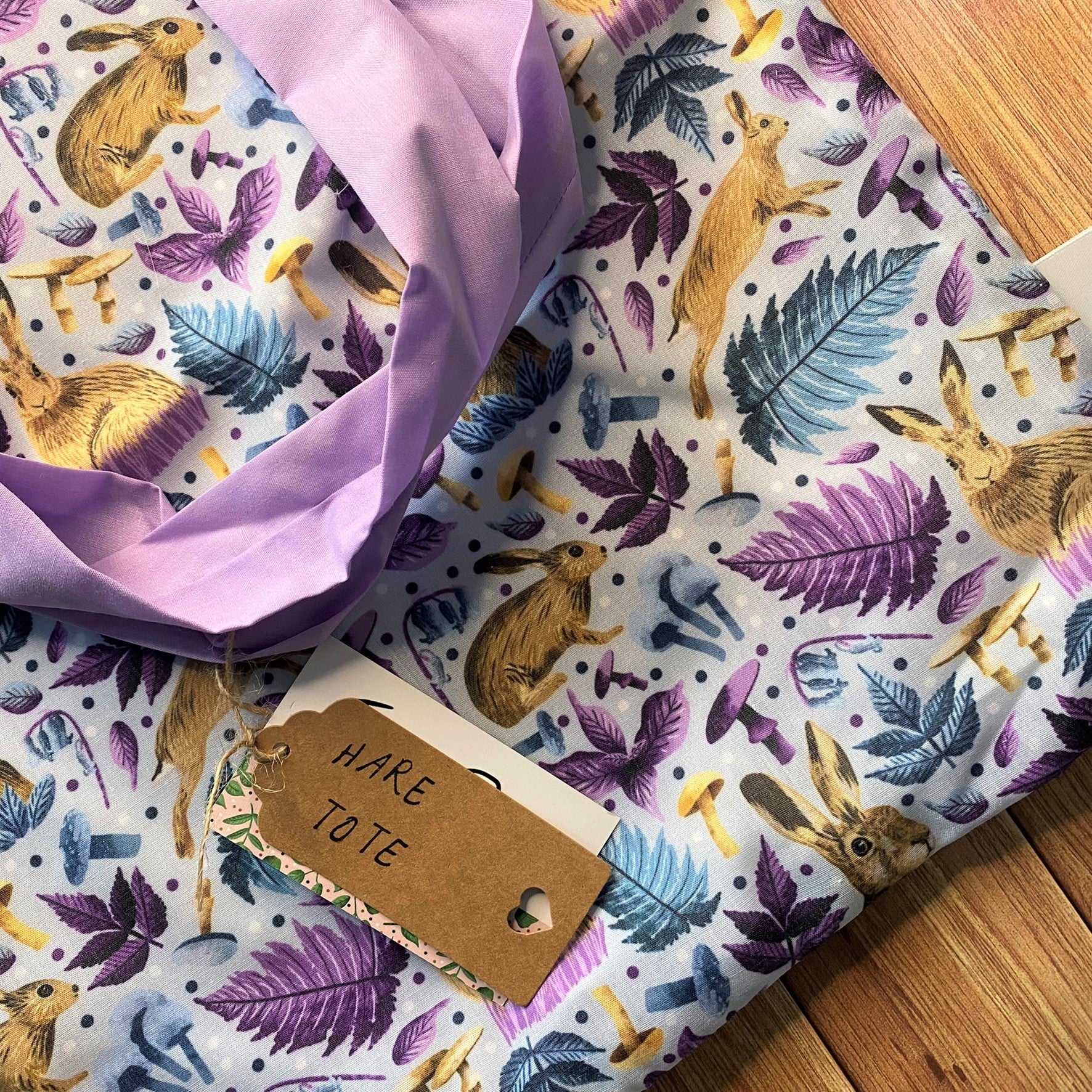 Closeup of surface pattern design on the tote bag featuring hares and foliage in purple and blue. It also shows a purple set of handles and the brown kraft label that says 'Hare Tote' on it.